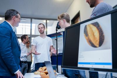 Student der Medieninformatik Lennaert Nitsche (Mitte) präsentiert dem Studiendekan Prof. Dr. Frank Thiesing (links) das Projekt seines Teams: Eine künstliche Intelligenz, die sechs Brötchensorten unterscheiden kann. (Foto: Hochschule Osnabrück / Elisa Stock)