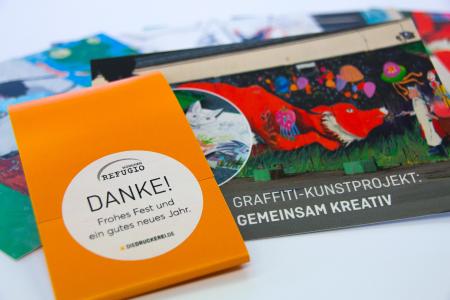 Die Jahresgabe 2017 für Sponsoren und Spender des Vereins Refugio München besteht aus sieben Postkarten mit Banderole und Aufkleber. Das Postkartenset zeigt Ausschnitte aus einem Wandbild, das im Rahmen des Kunstprojektes „on the road“ entstanden ist. Es ziert eine Hauswand der ehemaligen Bayernkaserne, heute eine Gemeinschaftsunterkunft für Geflüchtete. Produziert wurde das Postkartenset von diedruckerei.de / Copyright: Onlineprinters GmbH/ Sabine Mayer, Refugio München