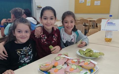 3 Mädchen sitzen am Frühstückstisch vor belegten Semmeln und lächeln.