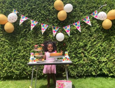 Fermosi steht im Garten hinter einem Tisch mit Kuchen, an der Hecke hinter ihr hängen Luftballons.