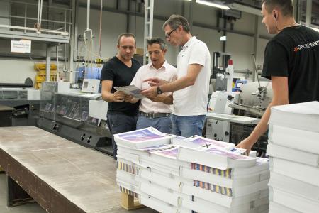 Als eine der ersten Druckereien in Europa wurde Onlineprinters auch im Bereich Druckweiterverarbeitung erfolgreich zertifiziert. Armin Schörghofer (2. v. r.), ugra-Auditor, begutachtet mit Co-Auditor Thomas M. Schnitzler Broschüren in der Weiterverarbeitung. Copyright: Onlineprinters GmbH
